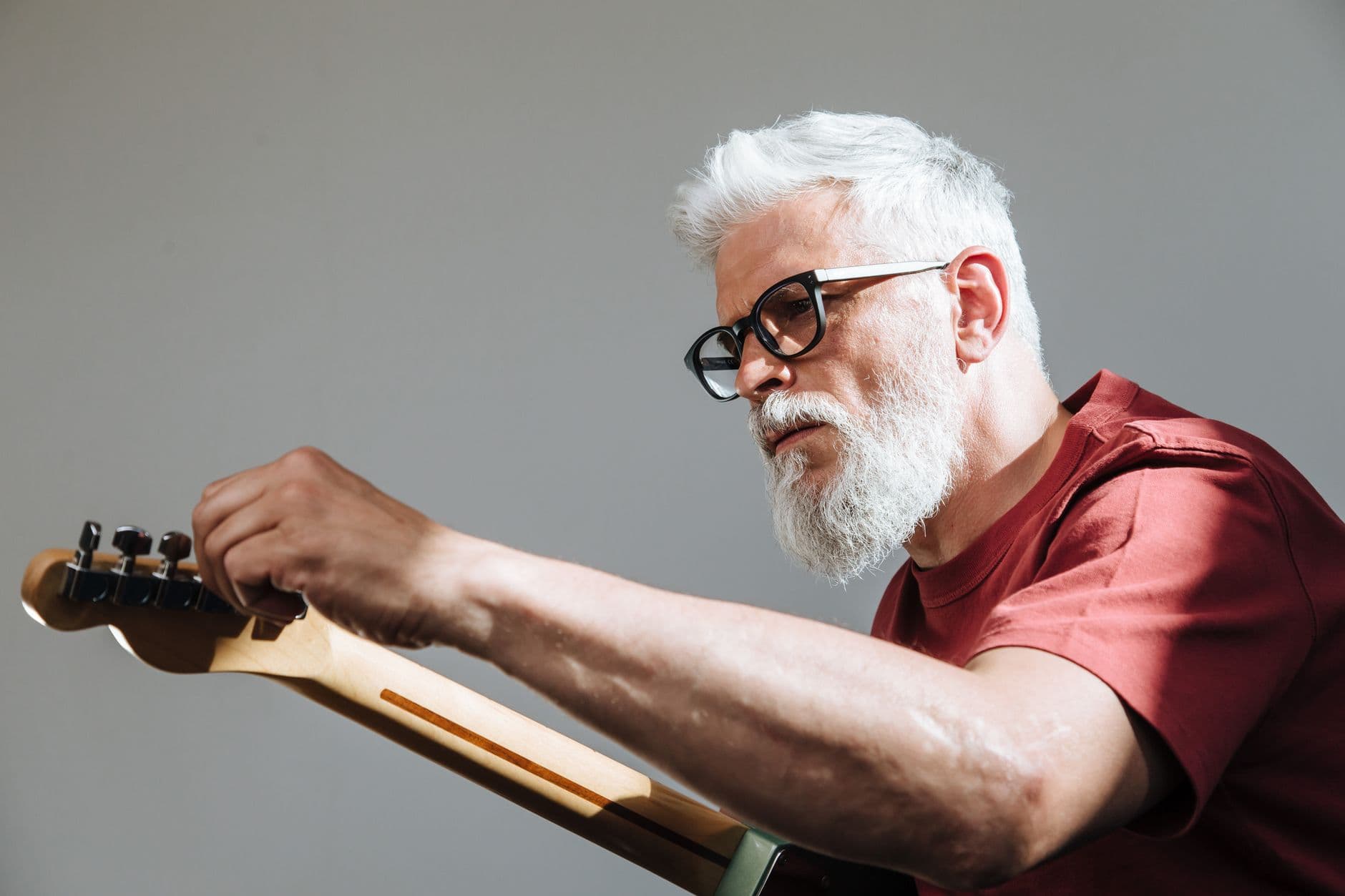A senior man with glasses adjusts an electric guitar indoors, showcasing focus and passion for music.