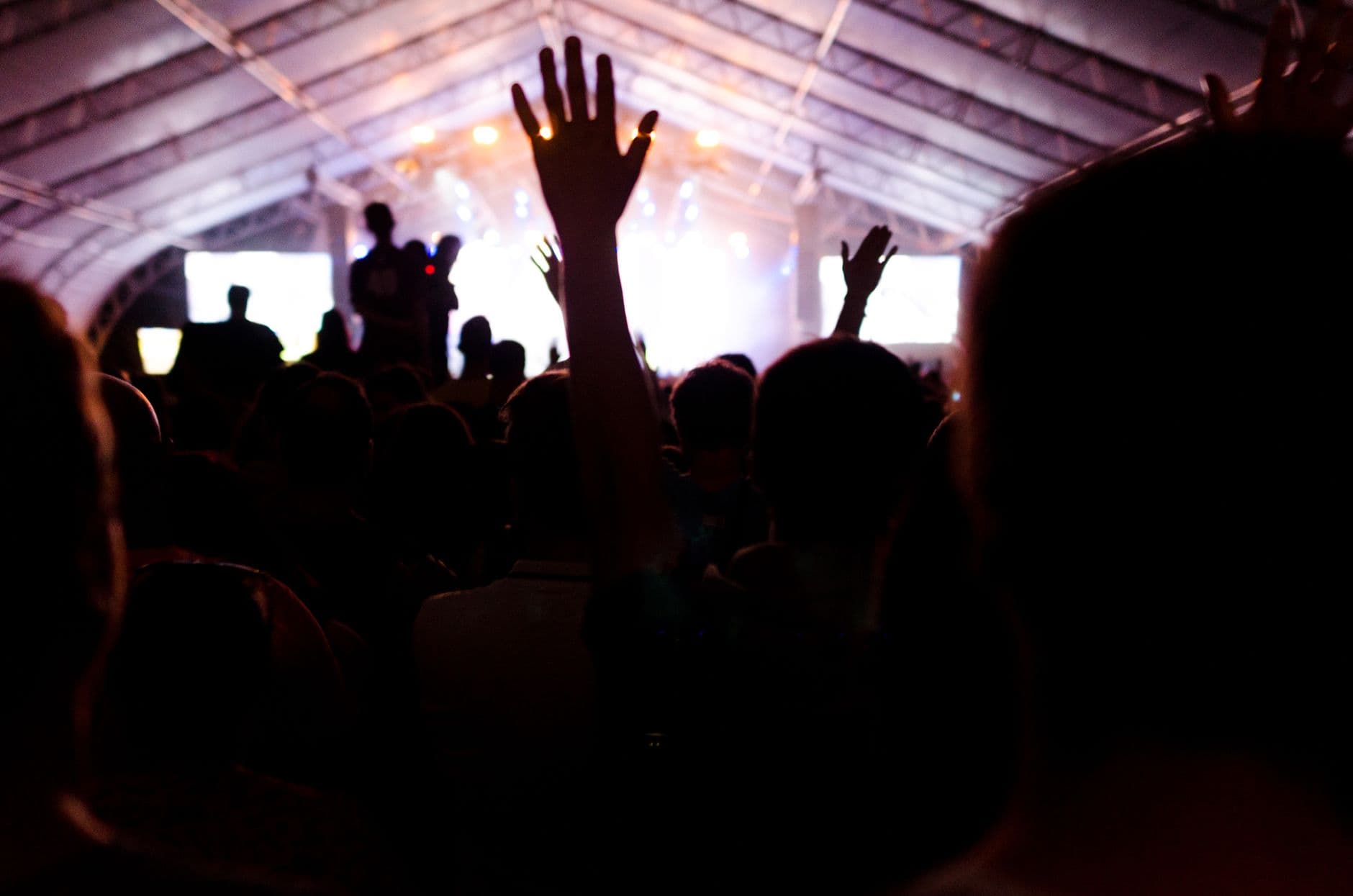 Energetic concert atmosphere with hands raised, vibrant lights, and a cheering crowd under a large tent.