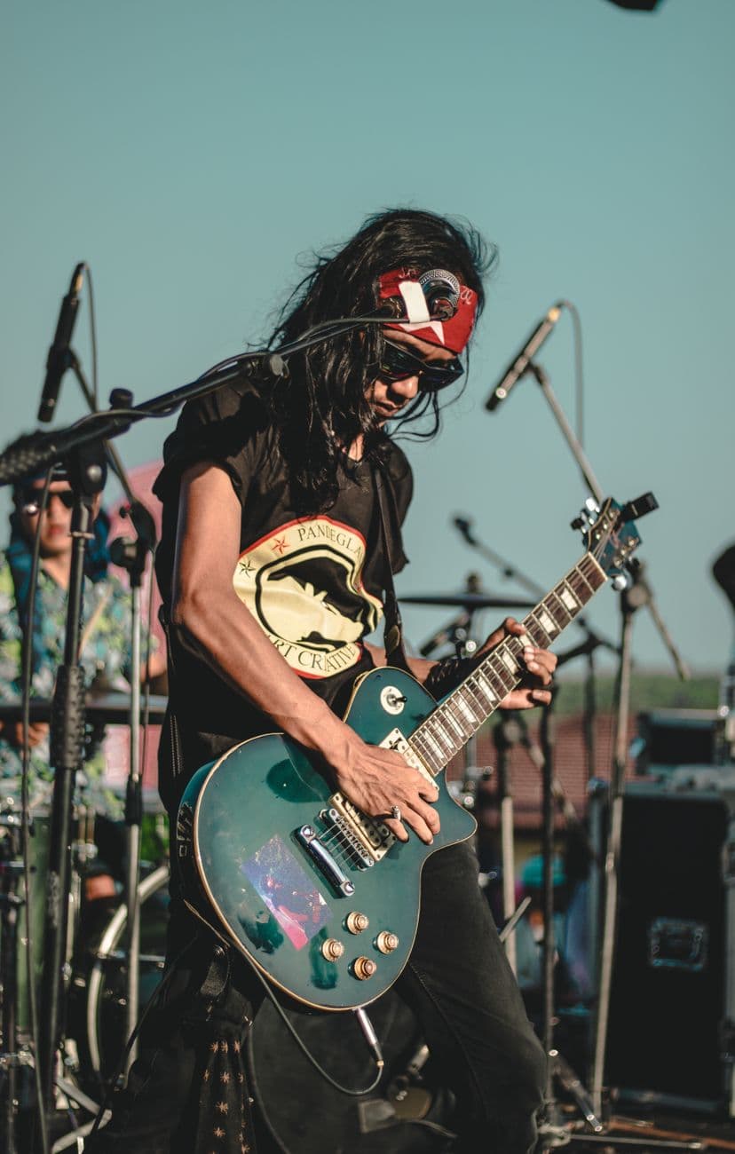 From below of ethnic male musician in bandana playing electric guitar near microphones on stage during performance