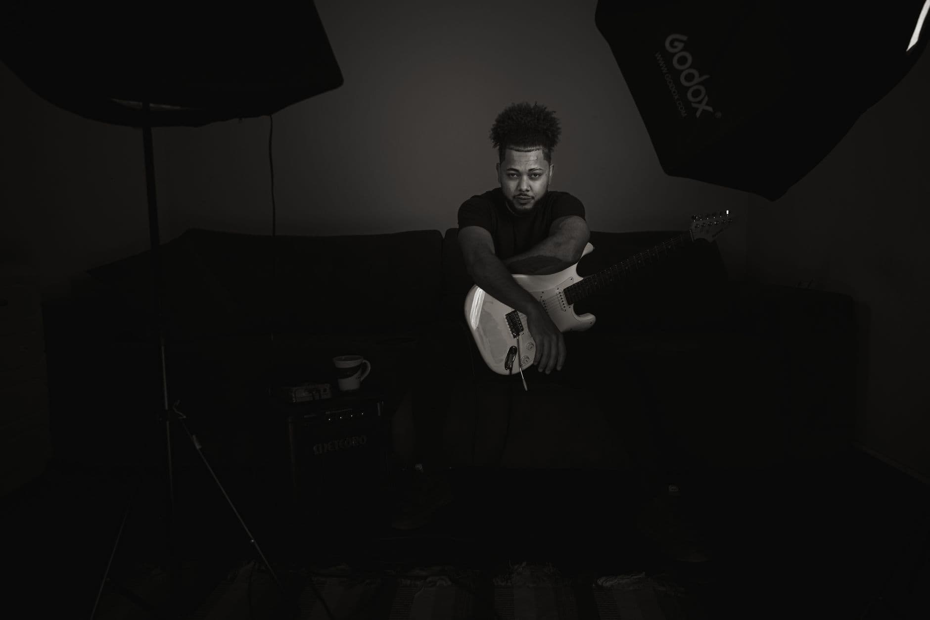 Black and white portrait of a man sitting on a couch holding an electric guitar in a moody setting.