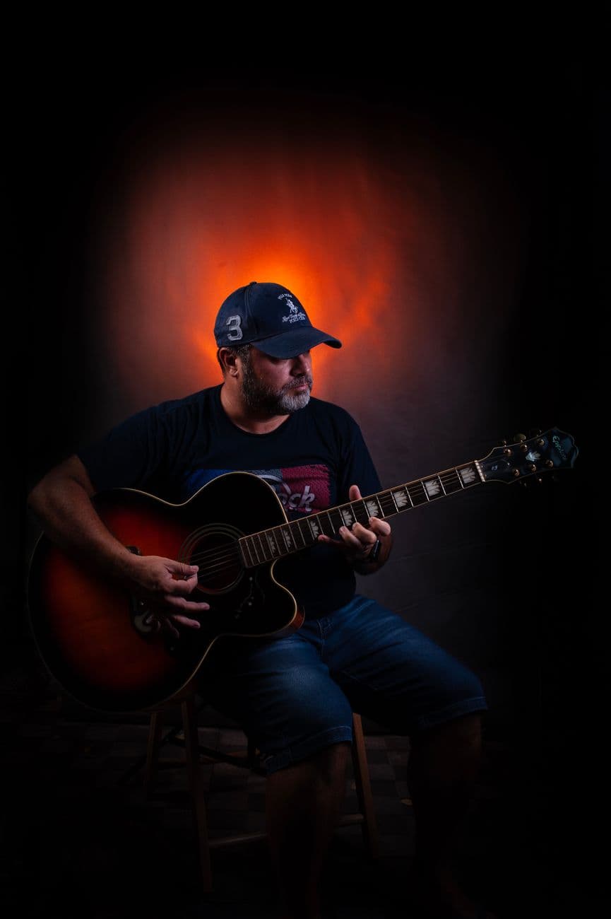Dreamy bearded male artist in cap playing acoustic guitar while sitting near bright glowing wall and looking away